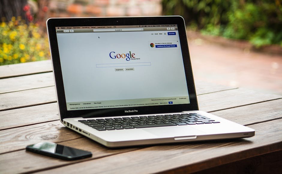 A MacBook Pro displaying Google Search on a wooden table outdoors, next to a smartphone. - Google SEO
